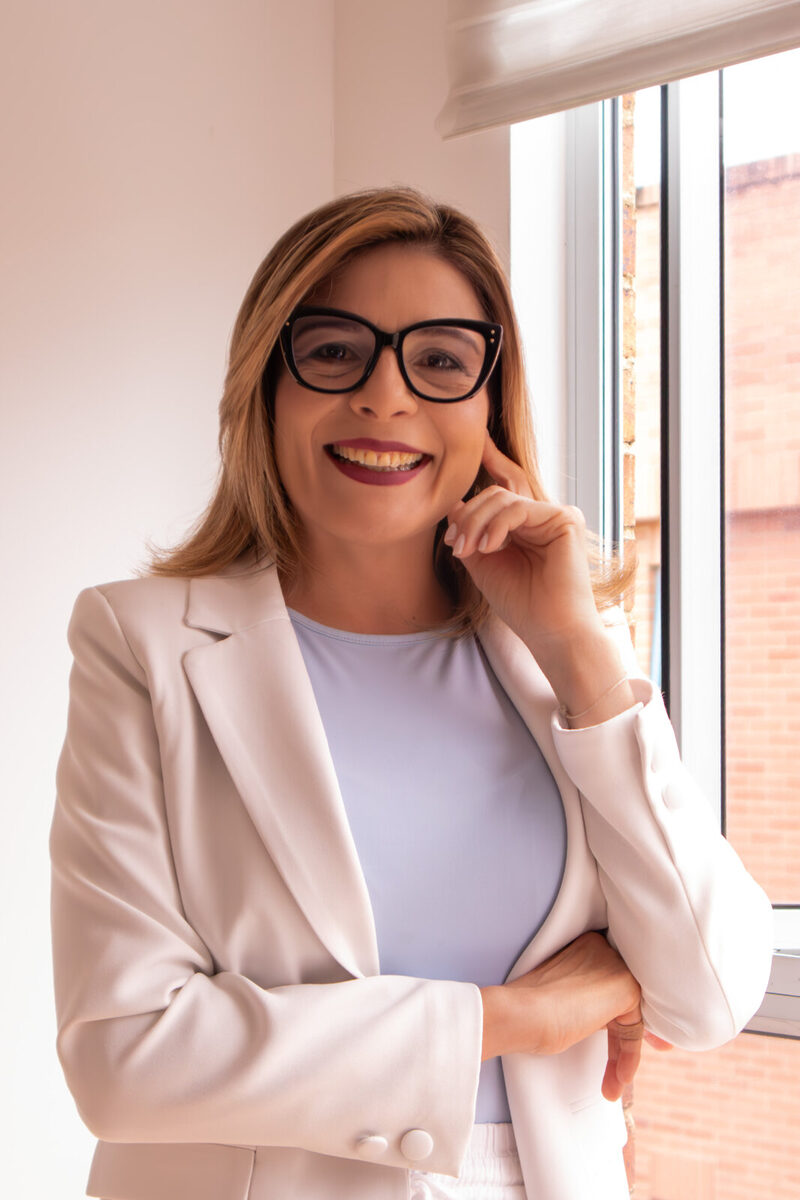 Retrato de Fabiola Quintero, sonriente, con blazer blanco y blusa lila, junto a la ventana de su consulta.